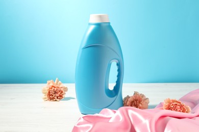 Photo of Bottle of fabric softener, clean cloth and carnation flowers on white wooden table against light blue background