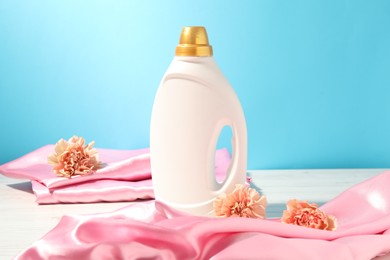 Photo of Bottle of fabric softener, clean clothes and carnation flowers on white wooden table against light blue background