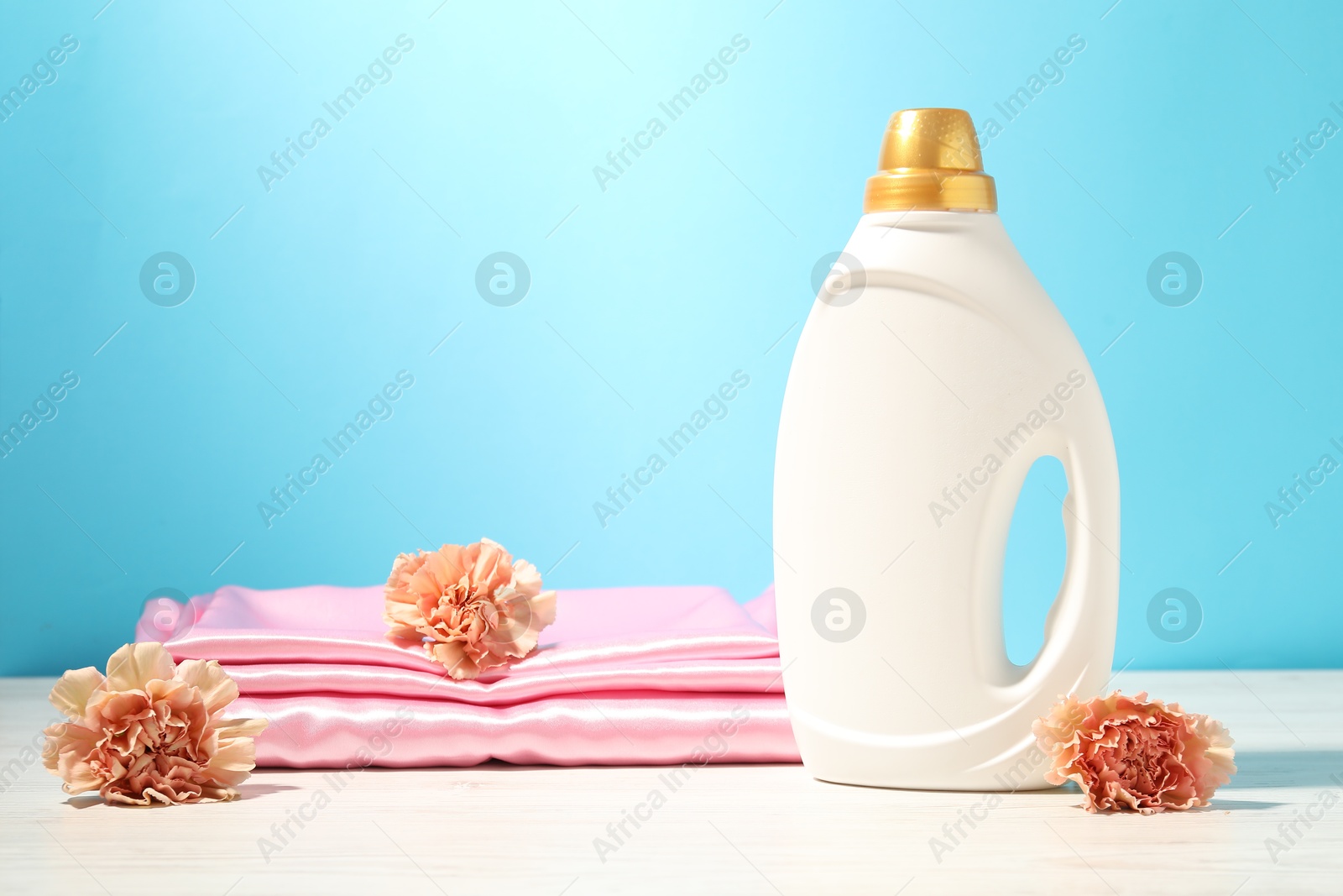 Bottle of fabric softener, clean clothes and carnation flowers on white wooden table against light blue background Photo of Bottle of fabric softener, clean clothes and carnation flowers on white wooden table against light blue background