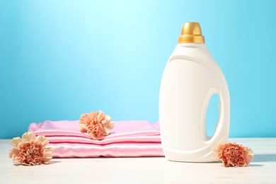 Photo of Bottle of fabric softener, clean clothes and carnation flowers on white wooden table against light blue background