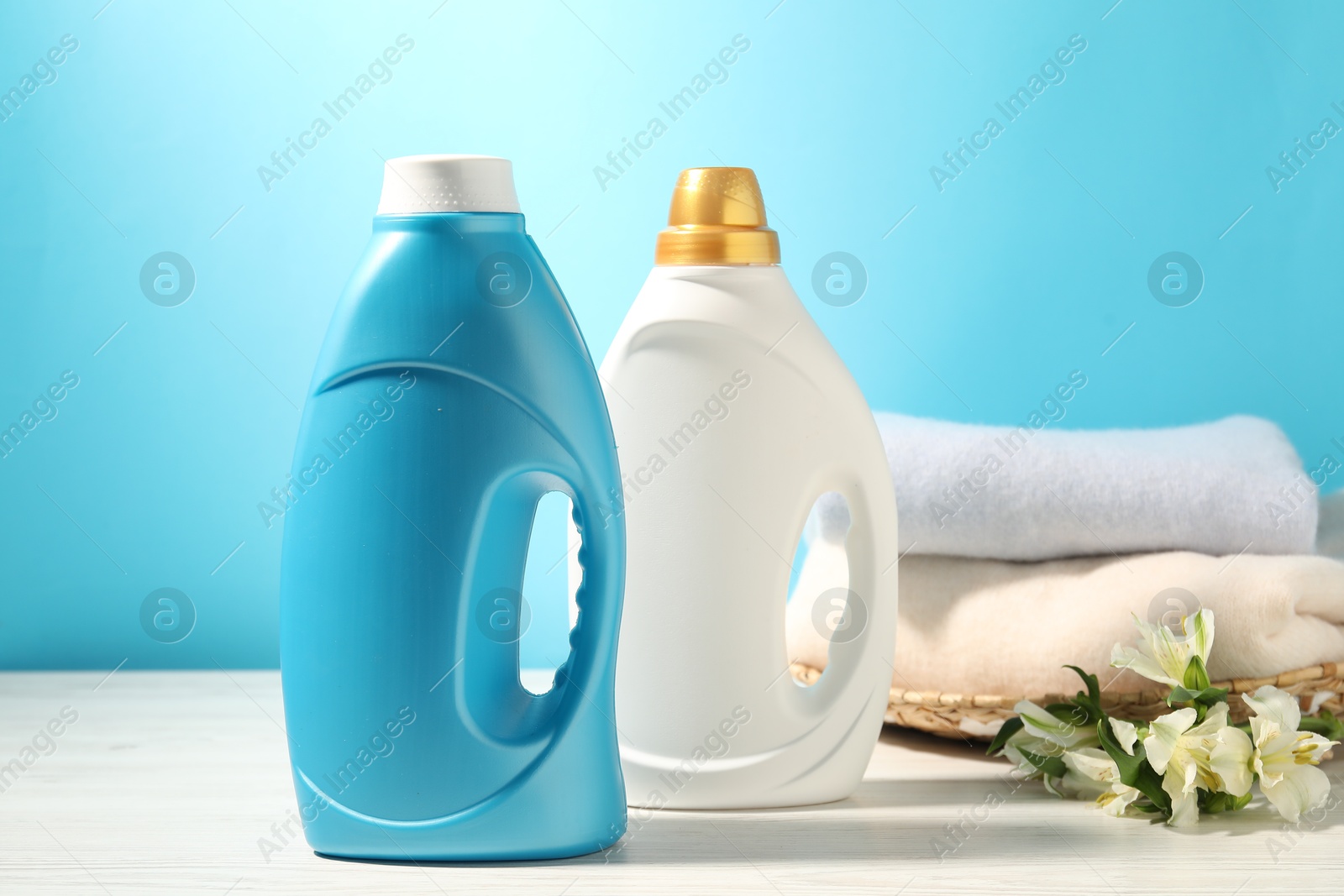 Photo of Bottles of fabric softener, clean clothes and lily flowers on white wooden table against light blue background