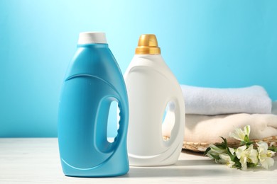 Photo of Bottles of fabric softener, clean clothes and lily flowers on white wooden table against light blue background