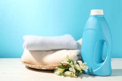 Photo of Bottle of fabric softener, clean clothes and lily flowers on white wooden table against light blue background