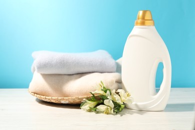 Photo of Bottle of fabric softener, clean clothes and lily flowers on white wooden table against light blue background