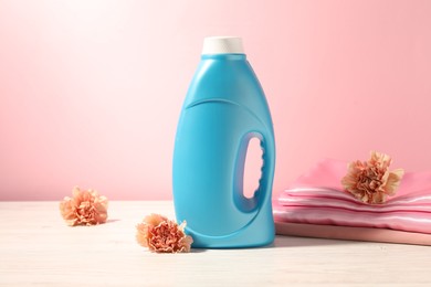 Photo of Bottle of fabric softener, clean clothes and carnation flowers on white wooden table against pink background