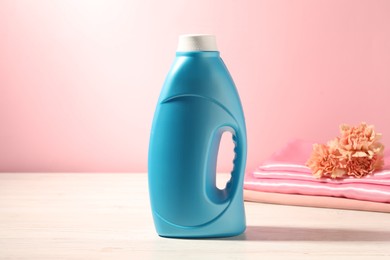 Photo of Bottle of fabric softener, clean clothes and carnation flowers on white wooden table against pink background