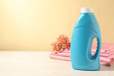 Photo of Bottle of fabric softener, clean clothes and carnation flowers on white wooden table against light yellow background. Space for text