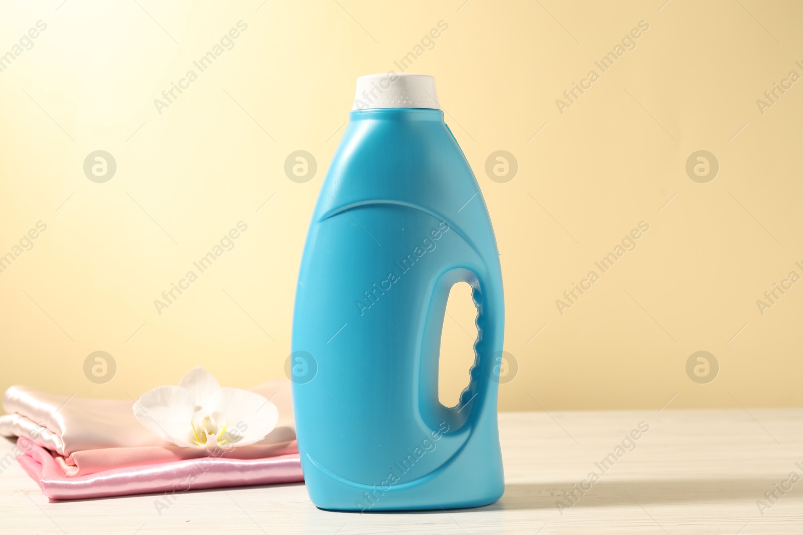 Bottle of fabric softener, clean clothes and orchid flower on white wooden table against light yellow background Photo of Bottle of fabric softener, clean clothes and orchid flower on white wooden table against light yellow background