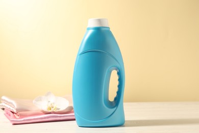 Photo of Bottle of fabric softener, clean clothes and orchid flower on white wooden table against light yellow background