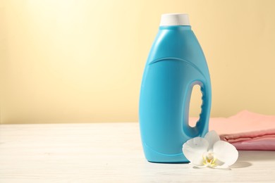 Photo of Bottle of fabric softener, clean clothes and orchid flower on white wooden table against light yellow background. Space for text
