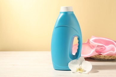 Photo of Bottle of fabric softener, clean clothes and orchid flower on white wooden table against light yellow background. Space for text