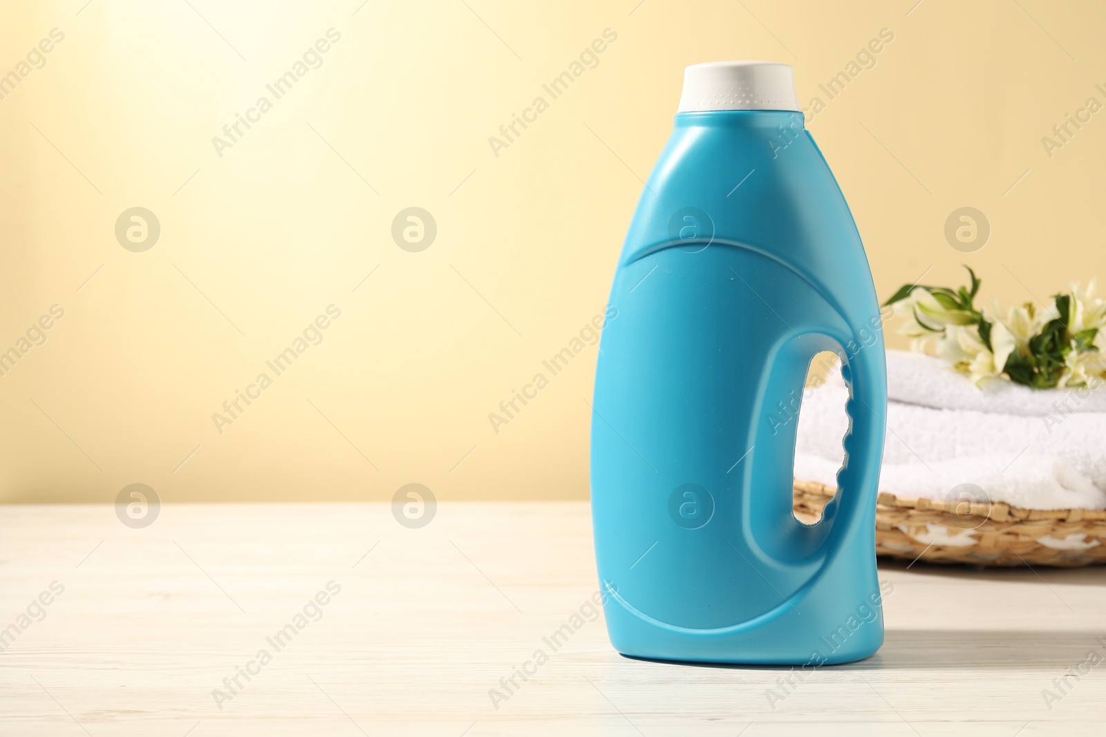 Photo of Bottle of fabric softener, terry towels and lily flowers on white wooden table against light yellow background. Space for text