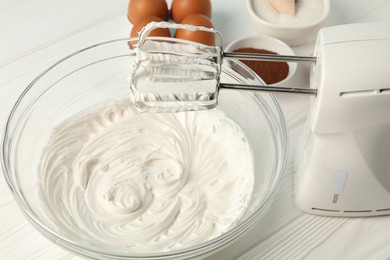 Photo of Bowl with whipped cream, hand mixer and ingredients on white wooden table, closeup