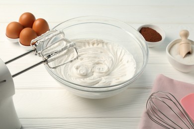 Photo of Bowl with whipped cream, hand mixer, whisk and ingredients on white wooden table, closeup