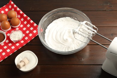 Photo of Bowl with whipped cream, hand mixer and ingredients on wooden table, closeup