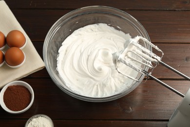 Photo of Bowl with whipped cream, hand mixer and ingredients on wooden table, flat lay
