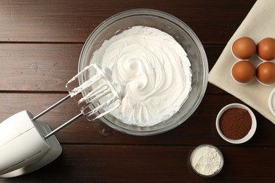 Photo of Bowl with whipped cream, hand mixer and ingredients on wooden table, flat lay