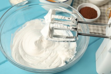 Photo of Bowl with whipped cream, hand mixer and ingredients on light blue wooden table, closeup