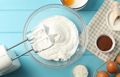Photo of Bowl with whipped cream, hand mixer and ingredients on light blue wooden table, flat lay