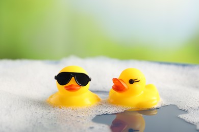 Photo of Cute rubber ducks floating in water with bubbles against blurred green background, closeup
