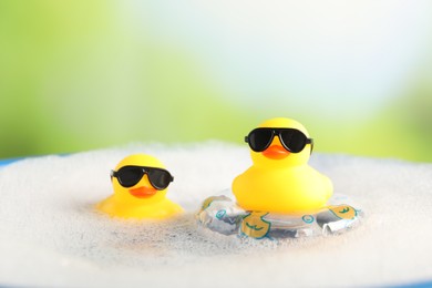 Photo of Cute rubber ducks floating in water with bubbles against blurred green background, closeup