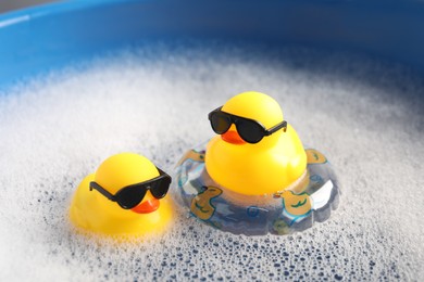 Photo of Cute rubber ducks floating in water with bubbles against blue background, closeup