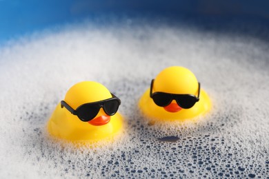 Photo of Cute rubber ducks floating in water with bubbles against blue background, closeup