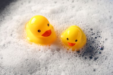 Photo of Cute rubber ducks floating in water with foam, closeup
