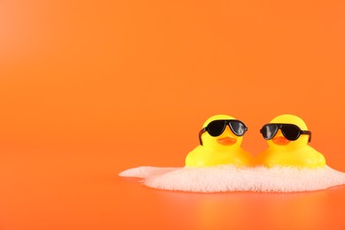 Photo of Cute rubber ducks with sunglasses and foam on orange background, space for text