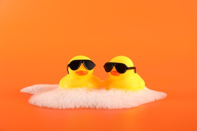 Photo of Cute rubber ducks with sunglasses and foam on orange background