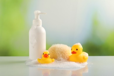Photo of Cute rubber ducks, foam, sponge and bottle of cosmetic product on table against blurred background