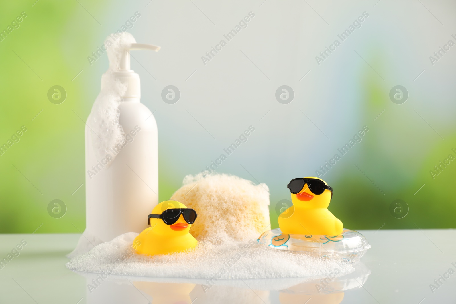 Cute rubber ducks, foam, sponge and bottle of cosmetic product on table against blurred background Photo of Cute rubber ducks, foam, sponge and bottle of cosmetic product on table against blurred background
