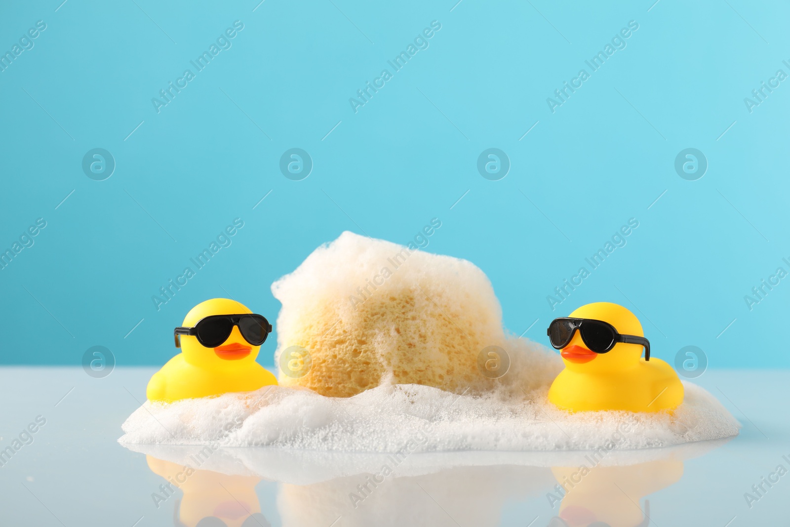 Cute rubber ducks, foam and sponge on table against light blue background Photo of Cute rubber ducks, foam and sponge on table against light blue background