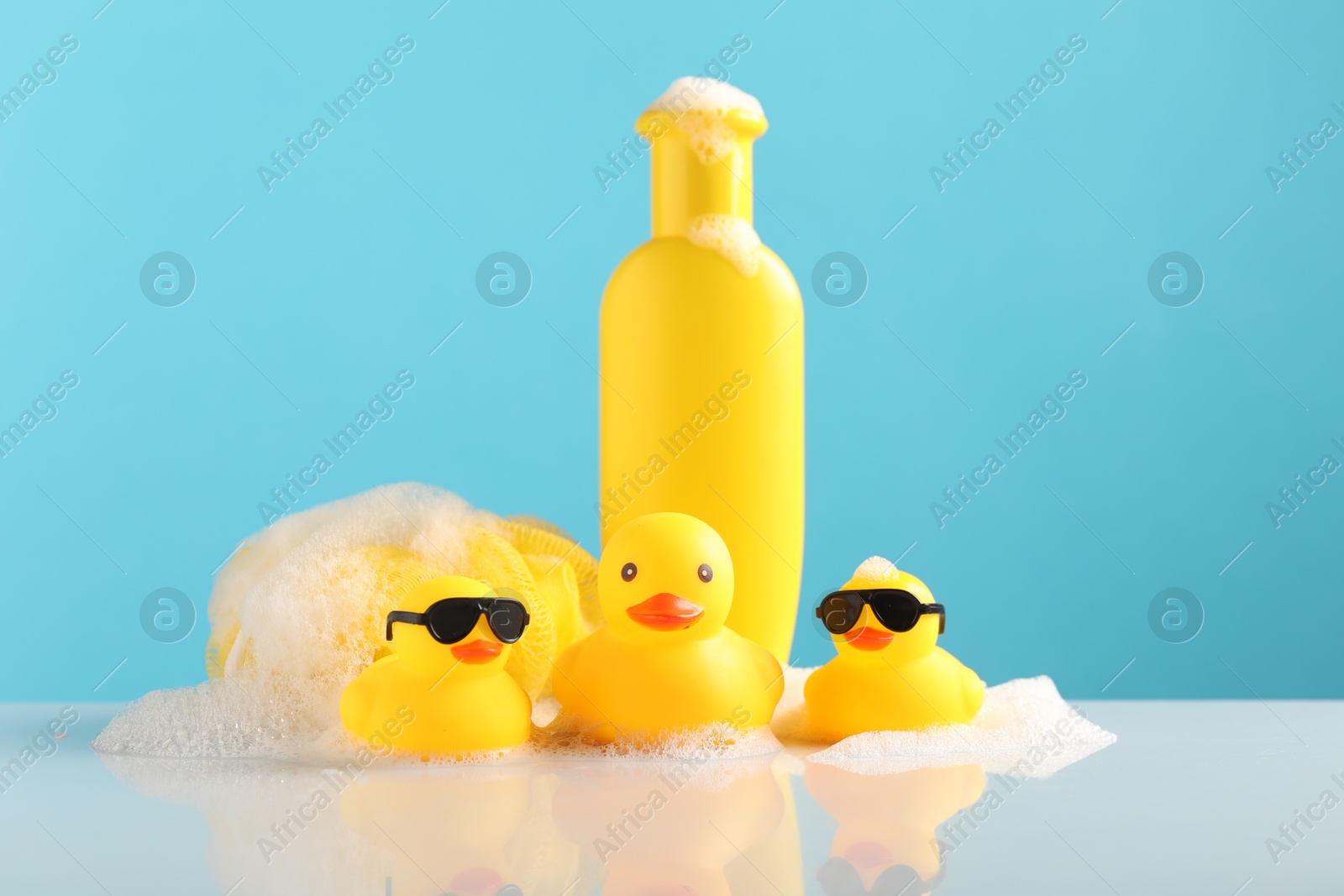 Cute rubber ducks, foam, sponge and bottle of shampoo on table against light blue background Photo of Cute rubber ducks, foam, sponge and bottle of shampoo on table against light blue background