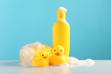 Photo of Cute rubber ducks, foam, sponge and bottle of shampoo on table against light blue background