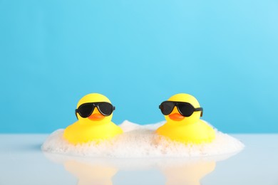 Photo of Two cute rubber ducks and foam on table against light blue background, closeup