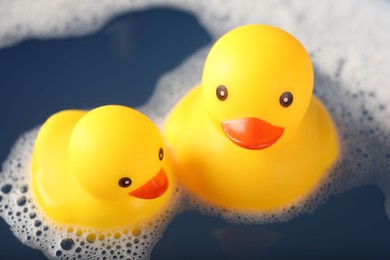 Photo of Cute rubber ducks floating in water with foam, closeup