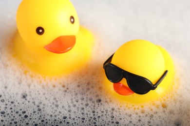 Photo of Cute rubber ducks floating in water with foam, closeup