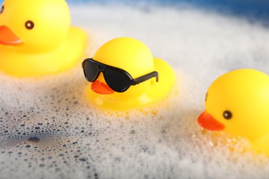 Photo of Cute rubber ducks floating in water with foam, closeup