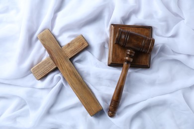 Photo of Judge's gavel and wooden cross on white fabric, flat lay