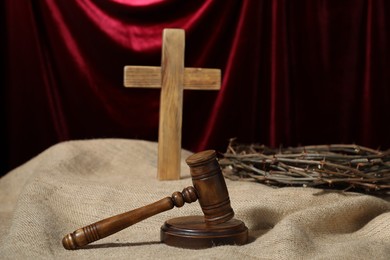 Photo of Judge's gavel, wooden cross and crown of thorns on burlap fabric against red curtain