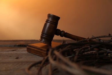 Photo of Judge's gavel and crown of thorns on wooden table against pale orange gradient background, closeup