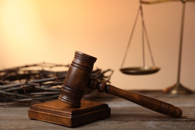 Photo of Judge's gavel, crown of thorns and scales on wooden table against pale orange gradient background, closeup