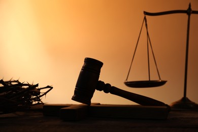 Photo of Judge's gavel, crown of thorns and scales on wooden table against pale orange gradient background, closeup