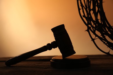 Photo of Silhouette of judge's gavel and crown of thorns on wooden table against pale orange gradient background, closeup