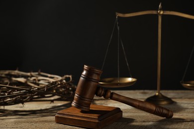 Photo of Judge's gavel, scales and crown of thorns on wooden table against black background, closeup