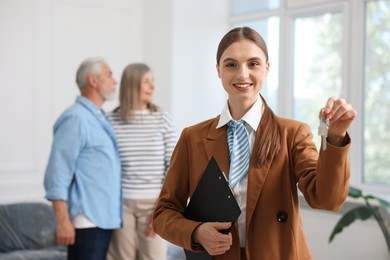 Photo of Smiling real estate agent with clipboard, house key and couple at home, selective focus
