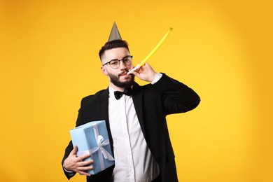 Photo of Happy Birthday. Handsome man with party hat, blower and gift box on orange background