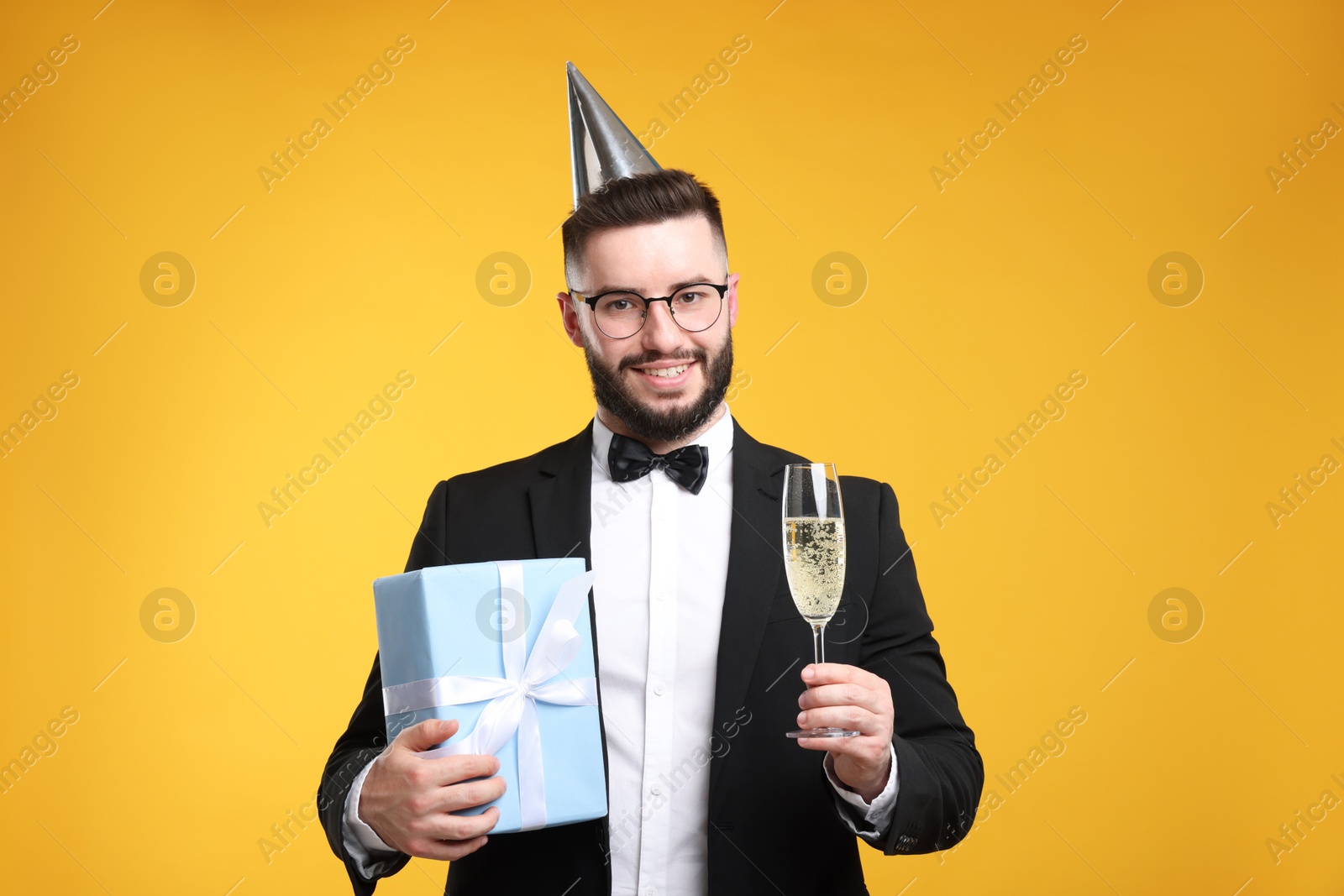 Happy Birthday. Handsome man with party hat, glass of champagne and gift box on orange background Photo of Happy Birthday. Handsome man with party hat, glass of champagne and gift box on orange background