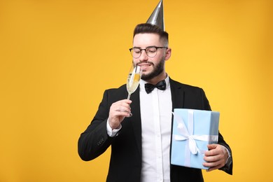 Photo of Happy Birthday. Handsome man with party hat, glass of champagne and gift box on orange background
